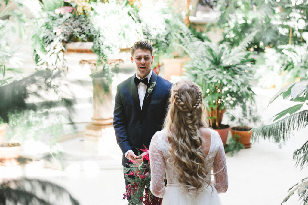 A groom looks at his bride, shown from behind. They are surrounded by plants.
